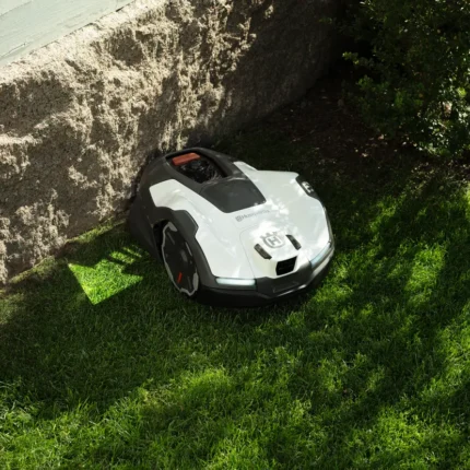 Robotic lawn mower operating on grass near a stone wall and bushes.
