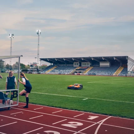 Ein Mähroboter, der auf einem Sportplatz in der Nähe einer Laufbahn arbeitet, mit einem Stadion im Hintergrund.