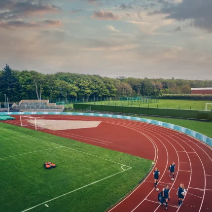 Ein Mähroboter, der auf einem Fußballfeld in der Nähe einer Laufbahn mit joggenden Menschen arbeitet.