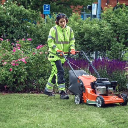 Eine Person in Arbeitskleidung, die einen Rasenmäher verwendet, um Gras in einer Gartenumgebung zu schneiden.