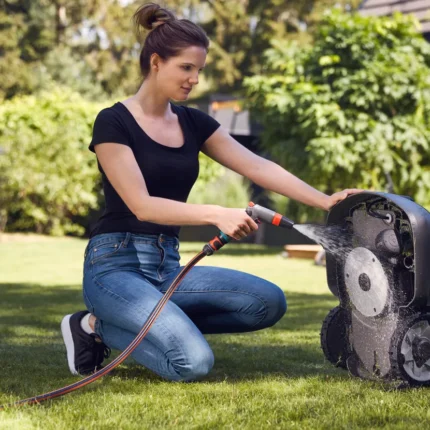 Eine Person, die einen Mähroboter in einem Hinterhof mit einem Gartenschlauch reinigt.