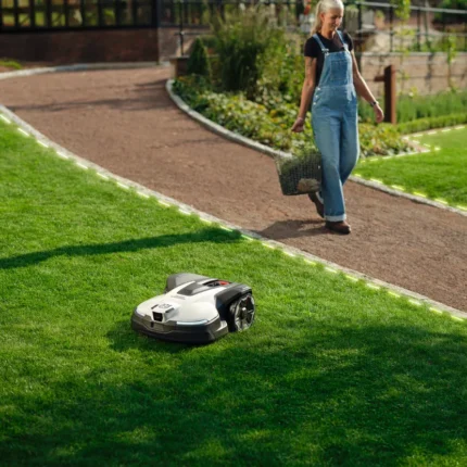 A person walking on a garden path with a robotic lawn mower operating on the grass.