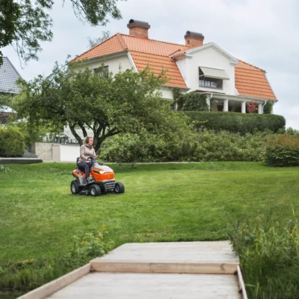 Eine Person, die mit einem Rider-Rasenmäher in einem Garten mit einem großen Haus im Hintergrund einen Rasen mäht.