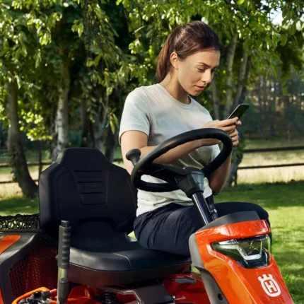 Eine Person, die auf einem Husqvarna Aufsitzmäher in einem Garten sitzt und ein Smartphone in der Hand hält.