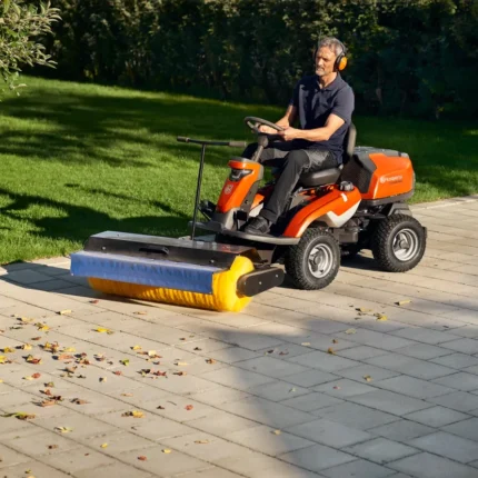 Eine Person, die eine Aufsitzkehrmaschine verwendet, um Blätter von einer gepflasterten Fläche neben einem Rasen zu entfernen.