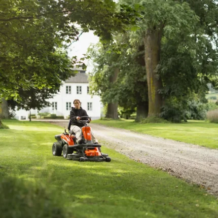 Eine Person, die mit einem Aufsitzmäher den Rasen in einer Gartenumgebung mit Bäumen und einem Haus im Hintergrund mäht.